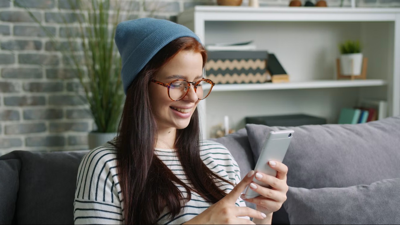 Junge Frau mit Mütze und Brille sitzt auf einem Sofa und liest lächelnd etwas auf ihrem Smartphone